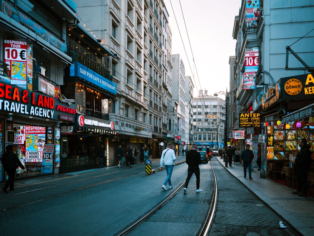 services-01 Vibrant street scene in Istanbul featuring shops, people, and tram tracks under a clear sky.