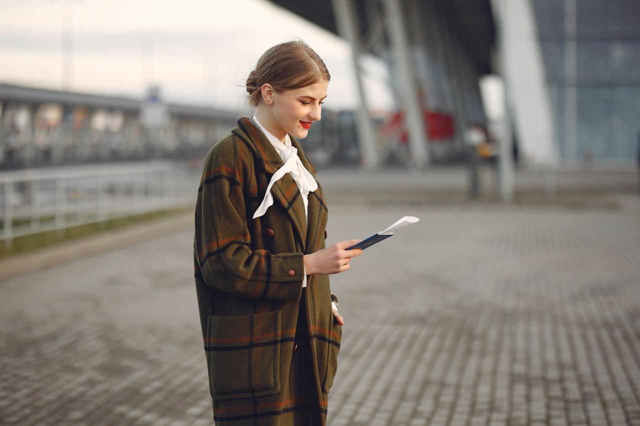 team-06 Smiling female passenger wearing trendy plaid coat and white blouse checking passport and ticket standing on pavement near contemporary building of airport