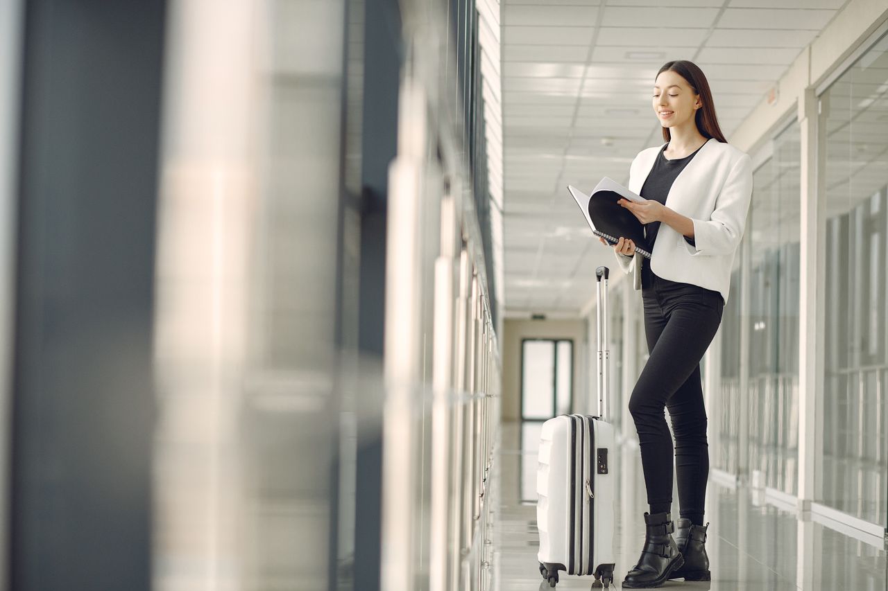 team-03 Full body of slim female businesswoman in formal clothes with suitcase standing in airport hallway and reading note in notebook while waiting for flight