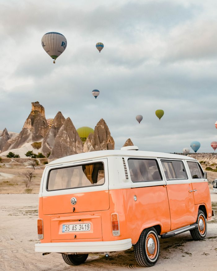 Vintage camper van parked on sandy ground near colorful air balloons floating in sky over  rocky terrain in Cappadocia