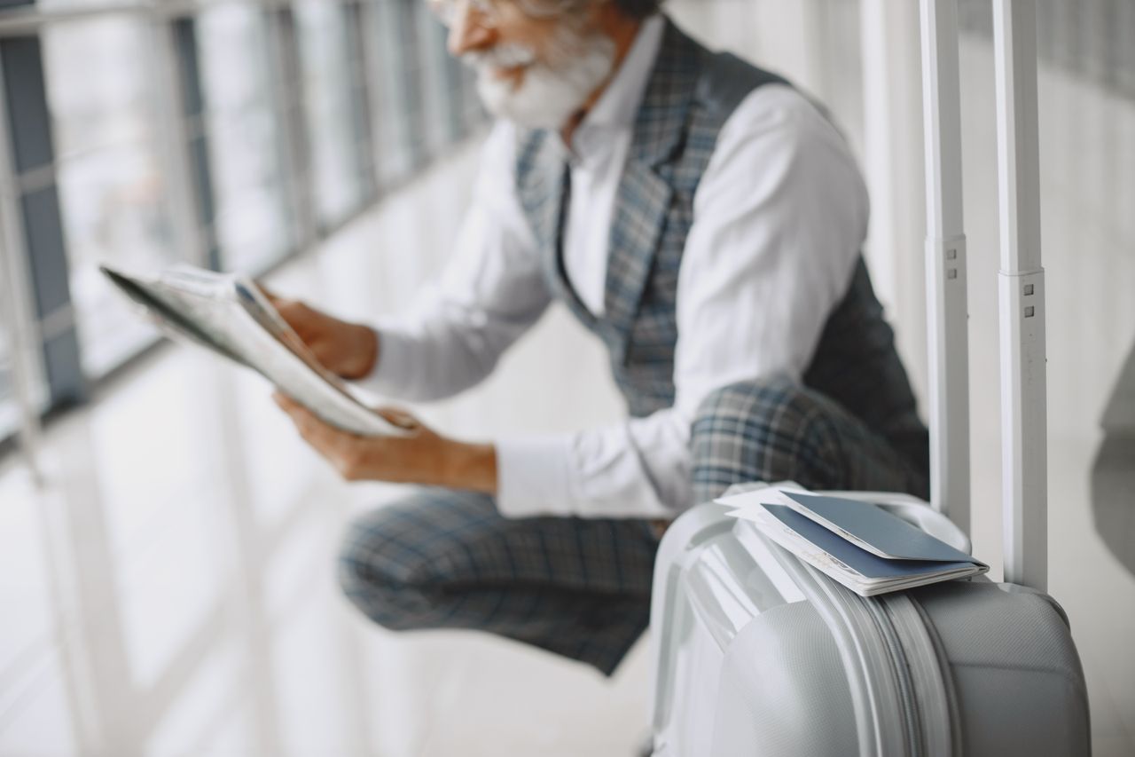 team-05 Adult man in plaid suit reads newspaper with suitcase at airport terminal.
